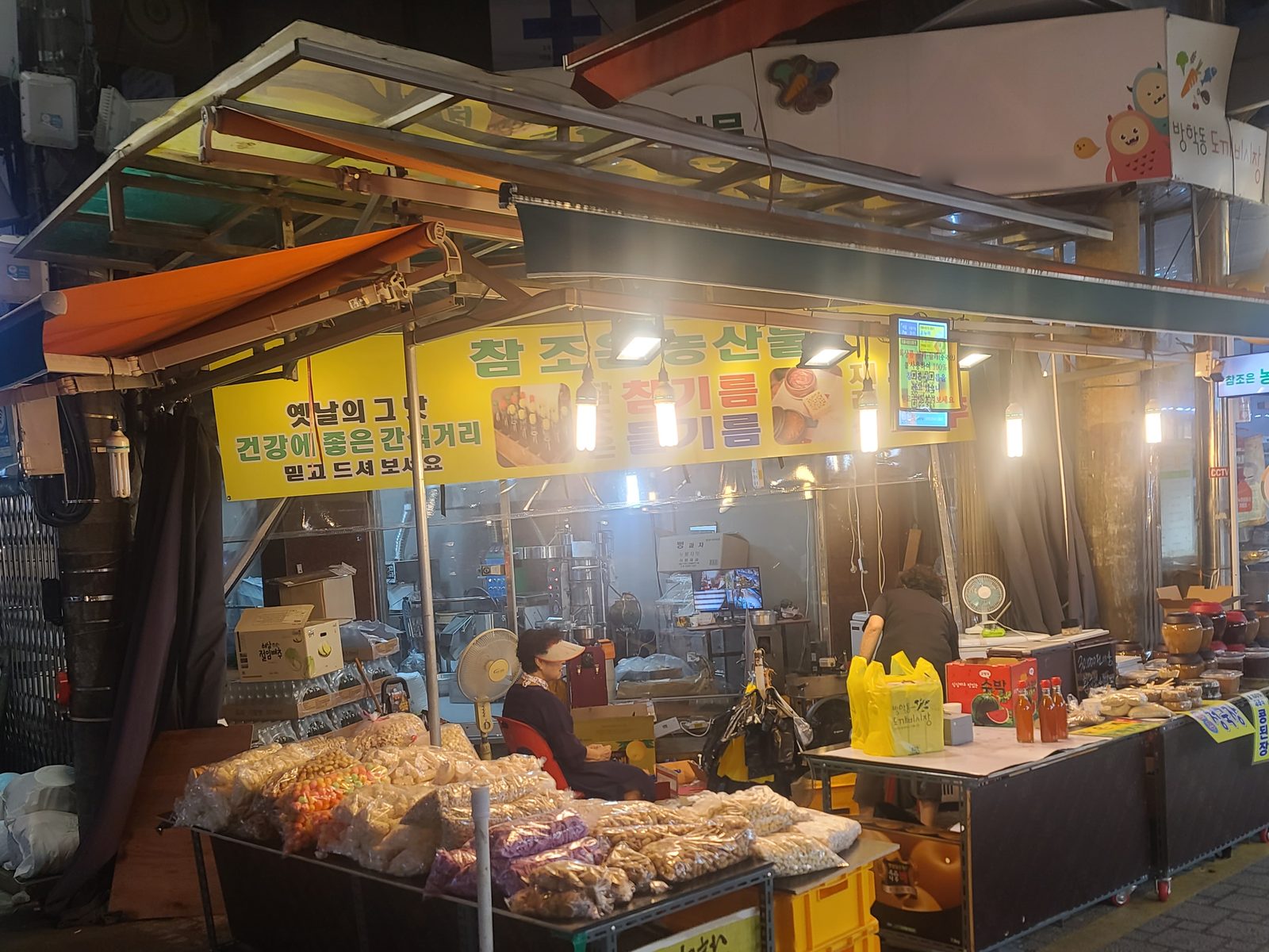 도봉구 방학동 도깨비시장 1문 참조은농산물 가게 전경. 노란 간판에 참기름·들기름이라 적혀 있고, 앞 매대에 강냉이와 뻥튀기 봉지가 놓여 있다.